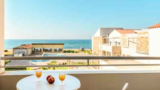 Utsikt fra en balkong med bord hvor to glass og to epler står, mot et basseng, bygninger og havet. (AI-generert tekst)