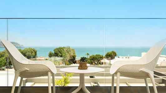 Utsikt fra en balkong med to hvite stoler og et bord med en liten plante, samt havet og grønne omgivelser i bakgrunnen. (AI-generert tekst)