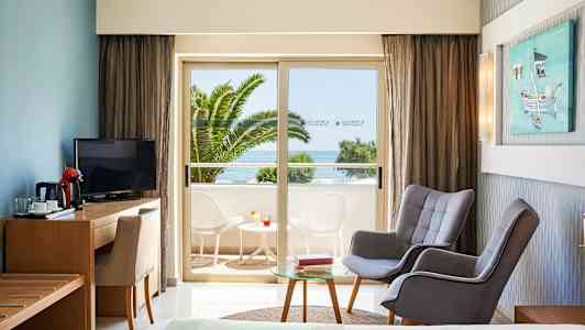 Hotellrom med sittegruppe og utsikt mot balkong med bord og stoler samt havutsikt. (AI-generert tekst)