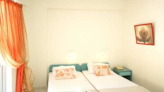 To enkeltsenger med oransje mønstrede puter, en telefon på et blått nattbord og oransje gardiner ved vinduet. (AI-generert tekst)