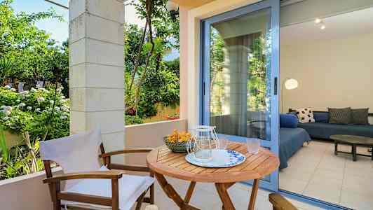 En utendørs terrasse med et trebord og stoler, omgitt av grønne planter. En glassdør fører til et rom med sittemøbler. (AI-generert tekst)