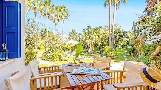 En terrasse med hagemøbler, utsikt over en solrik hage med palmer og vegetasjon. (AI-generert tekst)