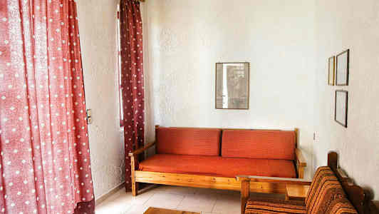 Stuen har røde gardiner, en tresofa med rødt rutete puter, og en stol med tekstiltrekk ved siden av. (AI-generert tekst)