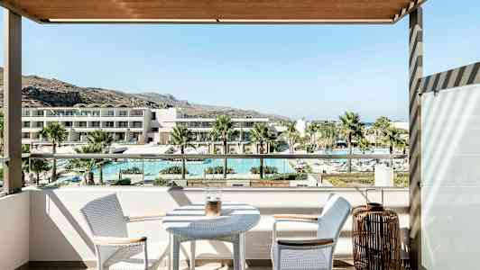 Utsikt fra en balkong med et bord og to stoler, som viser et basseng, palmer og moderne bygninger i bakgrunnen. (AI-generert tekst)