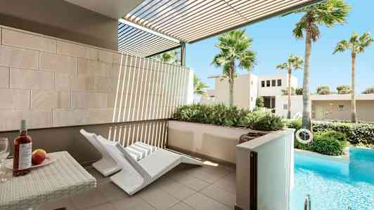 En utendørs terrasse med to hvite solsenger og et bord med vin og frukt. Foran terrassen er det et privat basseng og grønn vegetasjon. (AI-generert tekst)