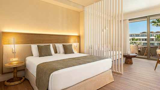 Et moderne hotellrom med en dobbeltseng, dekorative puter og et lite nattbord. Rommet har en skillevegg og en sittegruppe med utsikt over en balkong. (AI-generert tekst)