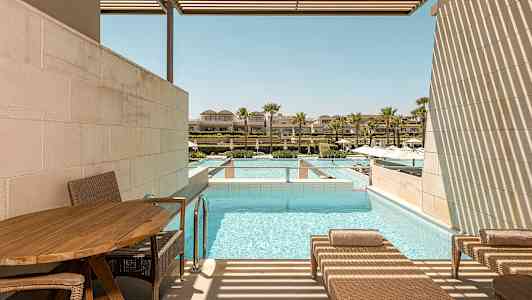 En uteplass med solsenger ved siden av et basseng. I bakgrunnen er det flere bygninger og palmer. (AI-generert tekst)