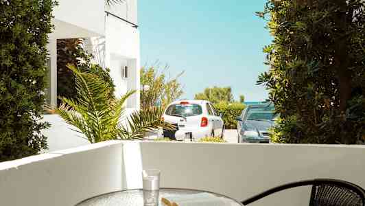 Terrasse med et bord hvor en bok og et glass står. Utenfor er det parkerte biler og vegetasjon. (AI-generert tekst)