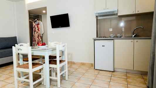 Et lite kjøkken med kjøkkenbenk, kjøleskap og spisebord med fire stoler, samt en sofa og veggmontert TV. (AI-generert tekst)