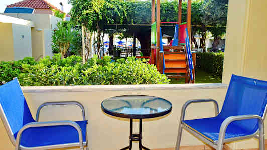 To blå stoler ved et bord med glasstopp på en terrasse, med lekeplass og grøntområde i bakgrunnen. (AI-generert tekst)