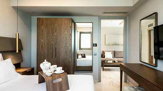 Et hotellrom med seng, tebrett, garderobe og speil. I bakgrunnen vises en stueområde med sofa. (AI-generert tekst)