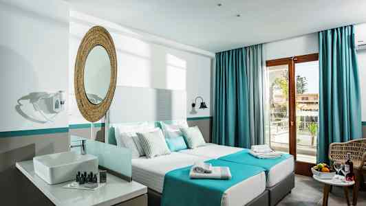 Et hotellrom med to enkeltsenger, et speil over en vask, og en balkongdør med blå gardiner. (AI-generert tekst)