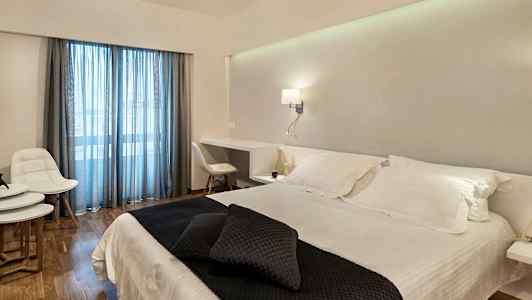 Et hotellrom med en dobbeltseng med hvite sengetøy og mørke pynteputer. Rommet har et skrivebord, hvite møbler og glassdører med blå gardiner. (AI-generert tekst)