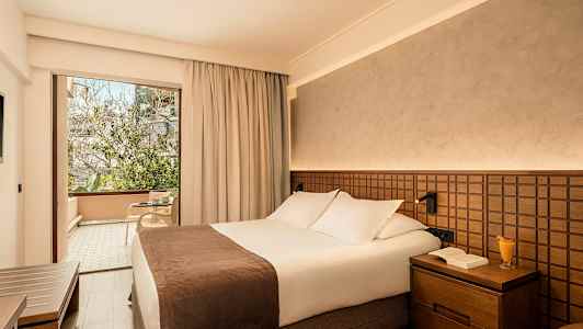 Hotellrom med en dobbeltseng og en glassdør som fører til en møblert balkong. (AI-generert tekst)