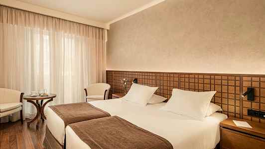 Et hotellrom med to enkeltsenger, brun innredning og en liten sittegruppe. (AI-generert tekst)