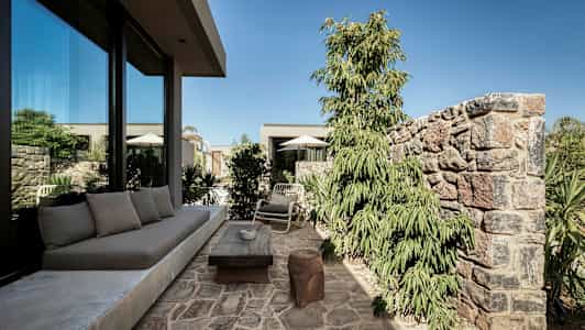 Steinlagt uteplass med sofa, bord og stoler nær et glasshus, omgitt av planter og murer. (AI-generert tekst)