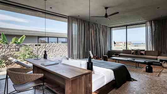 Moderne hotellsuite med stor seng, skrivebord og sitteområde. Vinduer med utsikt mot havet og vegetasjon. (AI-generert tekst)