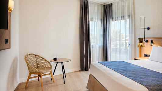 Et hotellrom med en moderne stol, et lite bord og en seng ved vinduer med lyse gardiner. (AI-generert tekst)