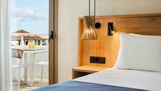 Hotellrom med en seng, treinnredning og en terrasse med bord og stoler i bakgrunnen. (AI-generert tekst)