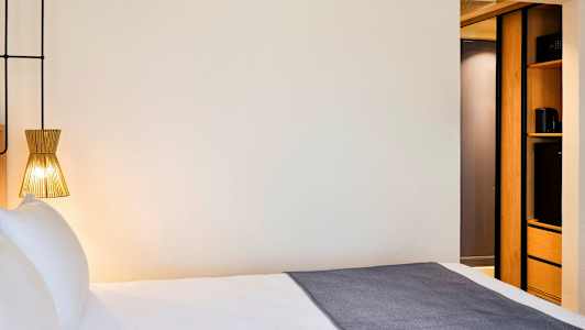 Et hotellrom med en seng, en moderne lampe og en del av innredningen med lagringsplass. (AI-generert tekst)