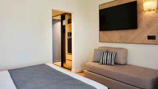 Et hotellrom med en seng, en sofa, en veggmontert TV og et åpent garderobeskap. (AI-generert tekst)