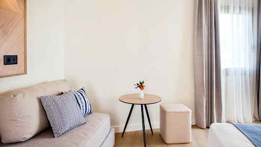 Et rom med en beige sofa, et trebord med en liten hvit vase og oransje blomster, en myk krakk og gardiner ved et vindu. (AI-generert tekst)