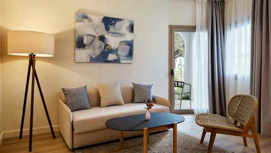 En stue med en sofa, et bord og en stol, samt en gulvlampe og gardiner foran vinduet. (AI-generert tekst)