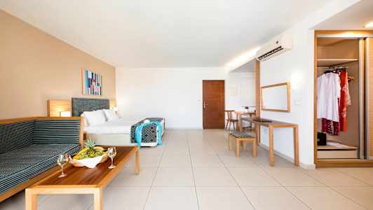 Et romslig hotellrom med dobbeltseng, sittegruppe, spisebord, speil og garderobe. En fruktskål og vinglass står på sofabordet. (AI-generert tekst)