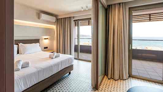 Hotellrom med seng og håndklær, store glassdører som fører ut til en balkong med havutsikt. (AI-generert tekst)