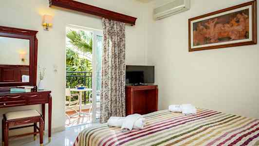 Et hotellrom med seng, TV, sminkbord og balkong med utsikt mot en palme. (AI-generert tekst)