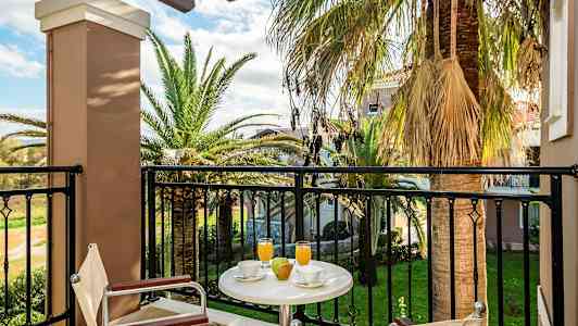 Utsikt fra en balkong med bord, stoler, drikke og frukt, samt palmer og hage i bakgrunnen. (AI-generert tekst)