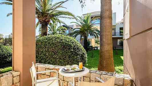 Utsikt fra en terrasse med bord og stoler mot en frodig hage med palmer og flerebolighus. (AI-generert tekst)