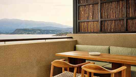 Terrasse med bord og stoler av tre, utsikt over havet og fjellene samt en vegg med bambus. (AI-generert tekst)