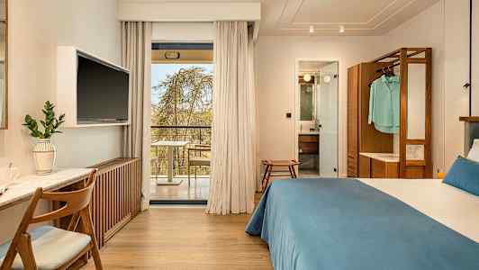 Hotellrom med dobbeltseng, skrivebord og TV. Glassdører fører ut til en balkong. Baderommet er synlig gjennom en åpen dør. (AI-generert tekst)