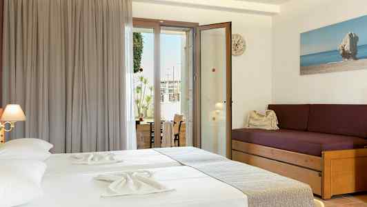 Et hotellrom med to senger med lyse laken, en lilla sofa med puter, og en glassdør som fører ut til en balkong. (AI-generert tekst)