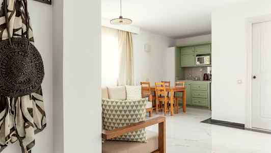 En stue med lys innredning, bord og stoler samt grønne kjøkkenskap. (AI-generert tekst)