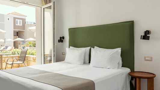 Hotellrom med en seng og en grønn sengegavl, lyse vegger og vindu med utsikt over en terrasse. (AI-generert tekst)