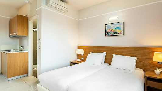 Hotellrom med to senger, nattbord og en liten kjøkkenkrok. (AI-generert tekst)