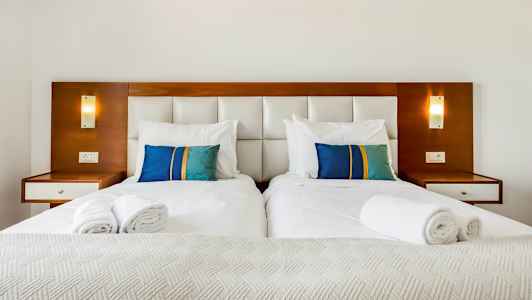 To enkeltsenger med hvite sengeklær og fargede puter i blått og grønt mot en sengegavl. (AI-generert tekst)