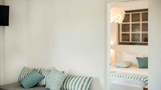 Et hotellrom med en sofa med puter i forgrunnen og en oppredd seng i bakgrunnen. (AI-generert tekst)