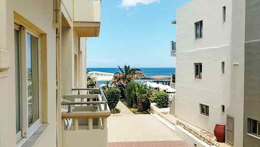 Bygninger med balkonger vender mot en gangsti omgitt av palmer, med havet i bakgrunnen. (AI-generert tekst)