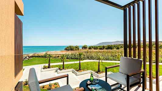 Bilde av en balkong med to sitteplasser, et bord med glass og avis, med utsikt mot en grønn hage og blått hav. (AI-generert tekst)