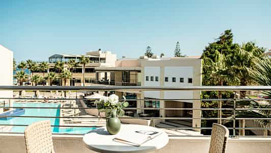 Utsikt fra en balkong med et bord, en stol og en vase med blomster, samt en åpen bok. I bakgrunnen ses en bygning og solområde med basseng. (AI-generert tekst)