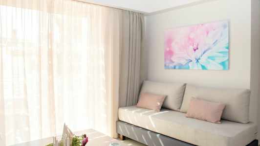 En lys og luftig stue med en beige sofa, rosa puter og et stort vindu med tynne gardiner. På veggen henger et blomsterbilde. (AI-generert tekst)