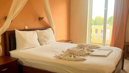 Et soverom med en dobbeltseng, hvitt sengetøy og en oransje vegg. Et stort vindu med gardiner ses i bakgrunnen. (AI-generert tekst)