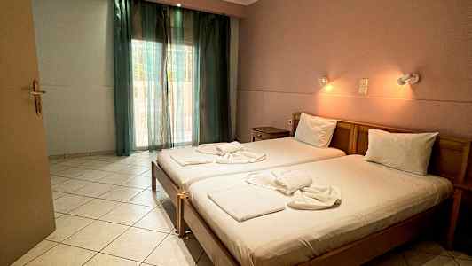 Et hotellrom med to enkeltsenger, oppredd og opplyst av vegglamper, samt et vindu med grønne gardiner. (AI-generert tekst)