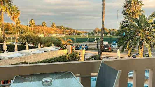 Utsikt fra en terrasse mot en idrettsplass omgitt av palmer og parkeringsplasser. (AI-generert tekst)