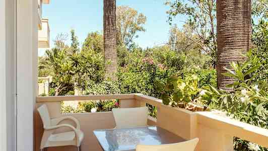 En balkong med bord og stoler, omgitt av grønne planter og palmer. (AI-generert tekst)