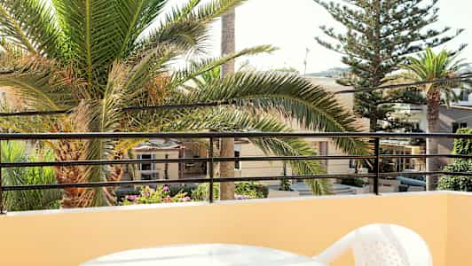 Utsikt fra balkong med et bord og plaststol, palmetrær og bygninger i bakgrunnen. (AI-generert tekst)