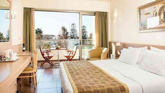 Hotellrom med dobbeltseng, skrivebord, sittemøbler og en balkong med to stoler og et bord. (AI-generert tekst)